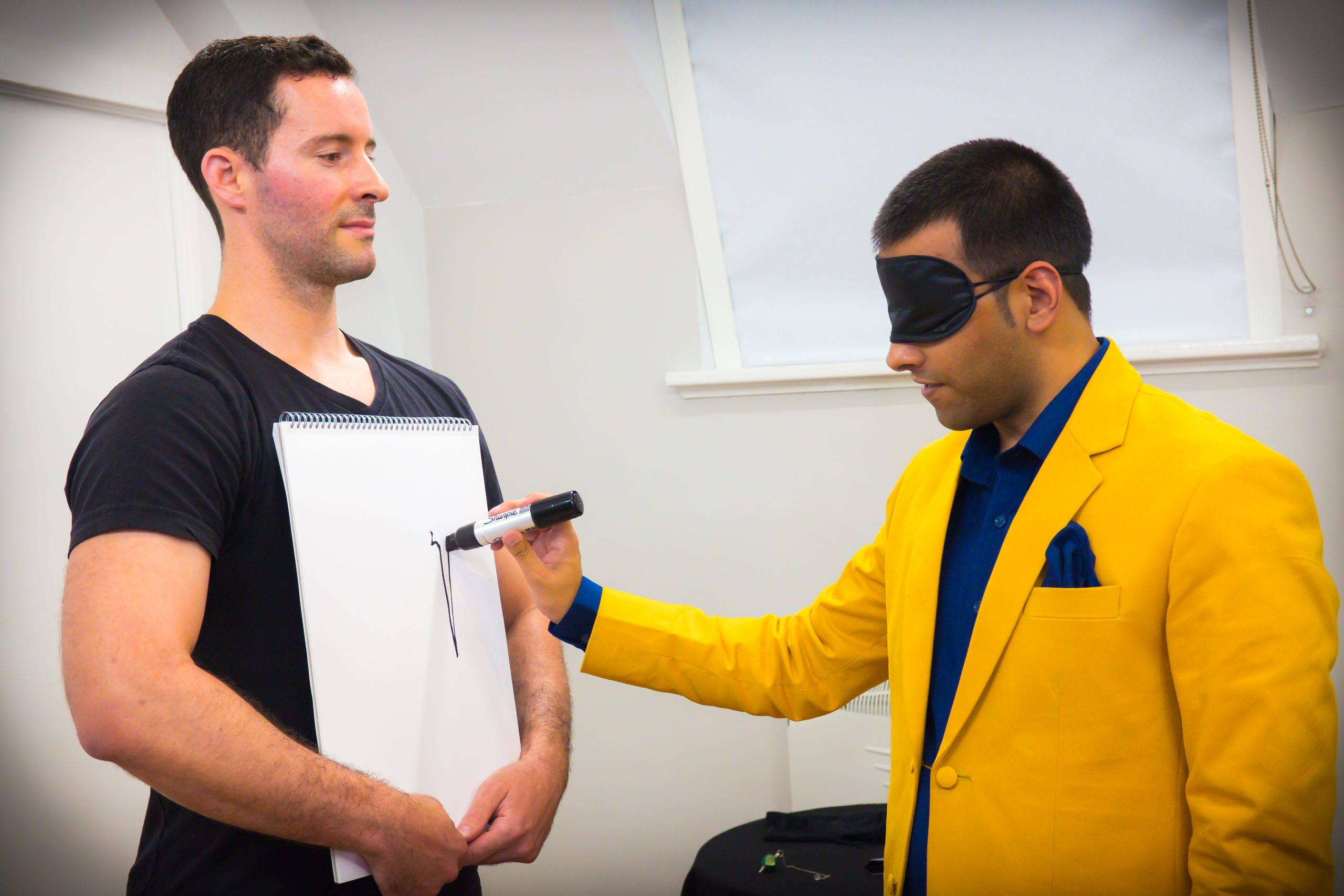 Drawing on a sketchpad whilst wearing a blindfold or sleep mask, dressed in a stylish yellow and blue bespoke suit, with a matching pocket square