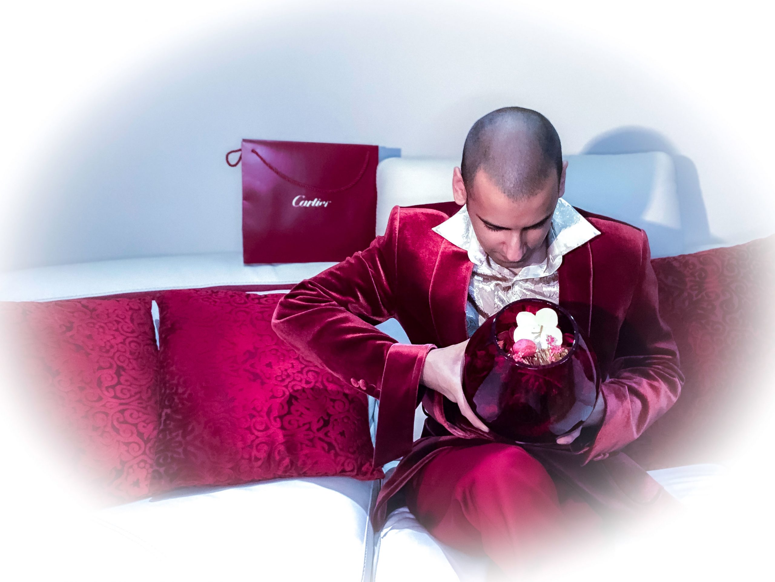 Dressed elegantly in a red robe and silk gold shirt whilst seated on a cosy cream living room sofa, holding a flowers vase with red cushions and a luxury designer bag from Cartier in the background