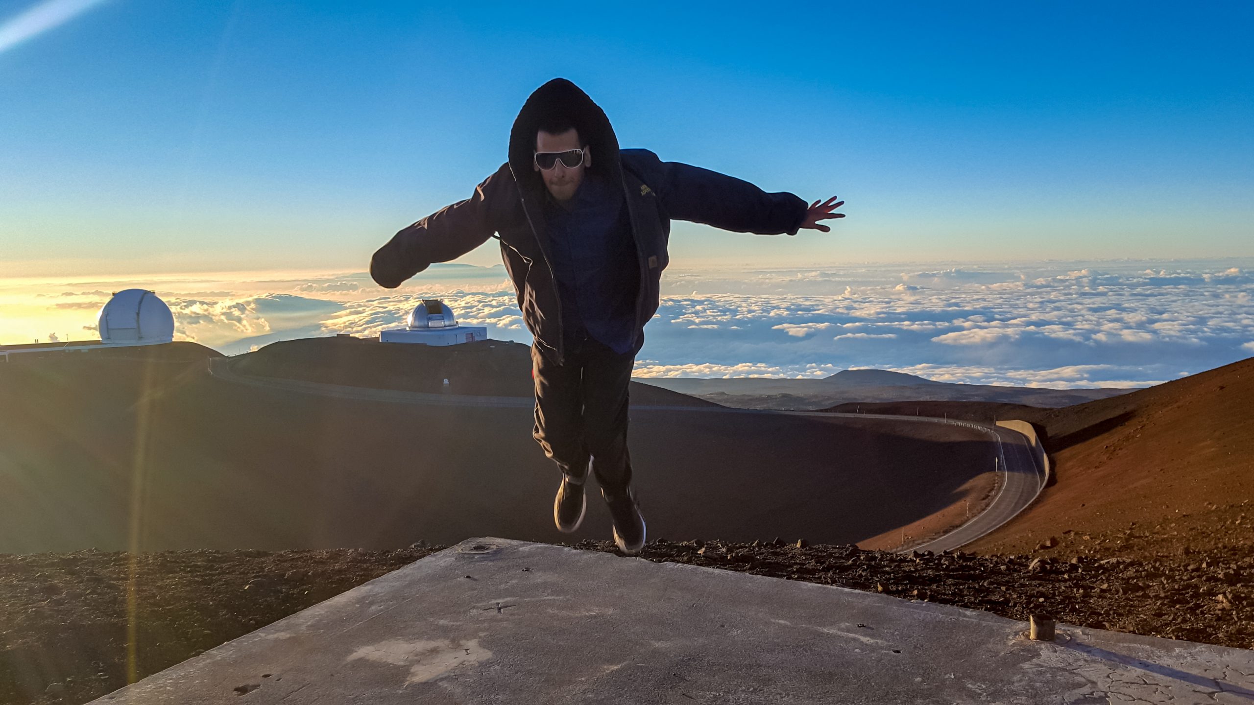 Girish jumping on a mountain in Hawaii, with clouds and a sunset in the background